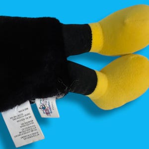 May include: A plush toy featuring black legs and yellow feet against a blue background. The toy has a tag attached. The feet are a bright yellow color, and the legs are black. The toy is made of soft material.