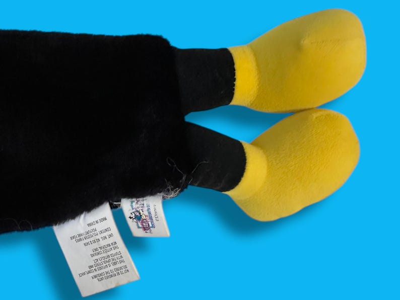 May include: A plush toy featuring black legs and yellow feet against a blue background. The toy has a tag attached. The feet are a bright yellow color, and the legs are black. The toy is made of soft material.