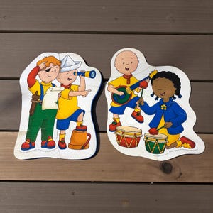 以下が含まれることがあります： 漫画のキャラクターが描かれた、2つのカラフルな木製ジグソーパズル。1つのパズルは海賊に扮した2人の子供を描き、もう1つは楽器を演奏する2人の子供を描いています。パズルは木製の表面にあります。