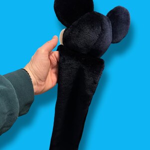 May include: A black plush Mickey Mouse head with large round ears and a long, black body. The plush toy has a white face and is held against a bright blue background.