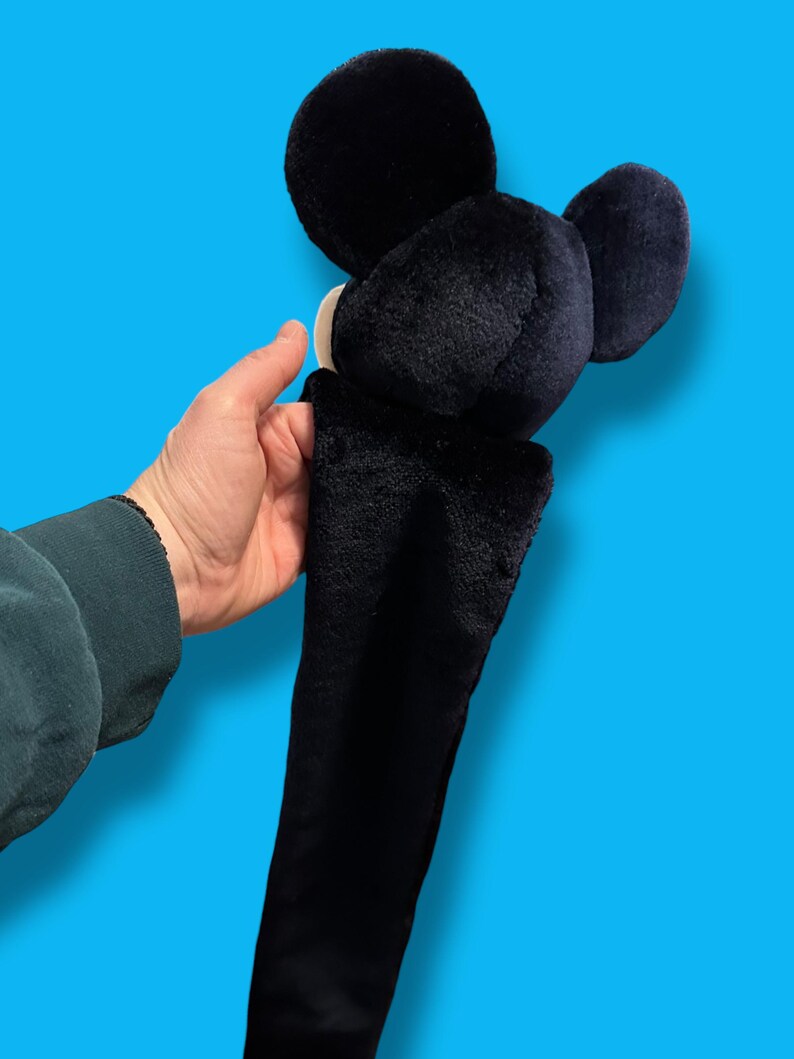May include: A black plush Mickey Mouse head with large round ears and a long, black body. The plush toy has a white face and is held against a bright blue background.