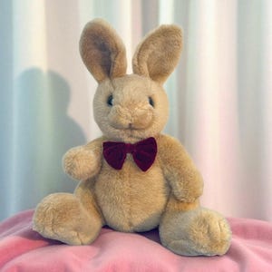 May include: A plush, tan-colored bunny rabbit with a burgundy bow tie. The stuffed animal has long ears, a smiling face, and is sitting on a pink surface. The bunny is a classic toy.