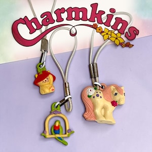 Peut inclure: Trois breloques Charmkins avec des cordons blancs. L'une est un chat jaune avec un chapeau rouge, une est un perroquet vert sur un perchoir jaune, et une est un poney crème avec une crinière et une queue roses, des taches violettes et une fleur blanche. Le texte Charmkins est visible.