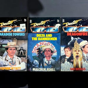 May include: Three vintage "Doctor Who" paperback books. Titles include "Paradise Towers," "Delta and the Bannermen," and "Dragonfire." Each cover features colorful illustrations and the Doctor Who logo. The books are displayed against a dark background.