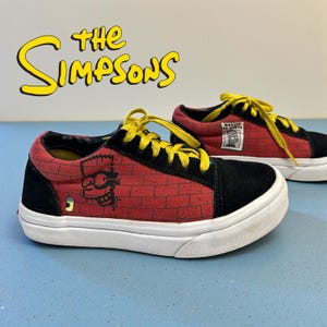 Puede incluir: Un par de zapatillas rojas y negras con cordones amarillos. Las zapatillas presentan un diseño de pared de ladrillos con gráficos de personajes de dibujos animados. El texto "THE SIMPSONS" se muestra encima de los zapatos.