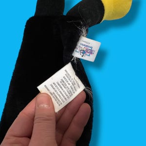 May include: A black plush toy with a yellow beak and head, possibly a toucan or similar bird. The toy has a tag with text in English and French. The background is a solid blue.