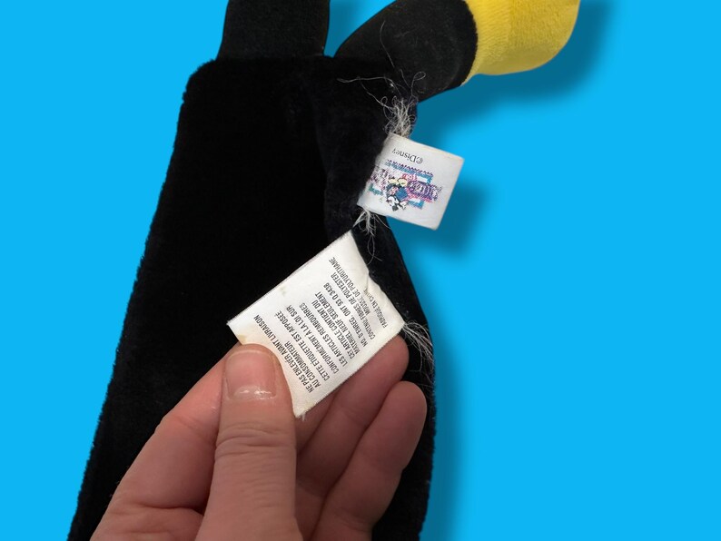 May include: A black plush toy with a yellow beak and head, possibly a toucan or similar bird. The toy has a tag with text in English and French. The background is a solid blue.