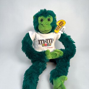 Peut inclure: Une peluche singe avec une fourrure vert foncé et un visage, des mains et des pieds vert clair. Le singe porte un t-shirt blanc avec le logo M&M's World et le mot Orlando en rouge. Une étiquette jaune M&M's World est attachée à l'oreille.