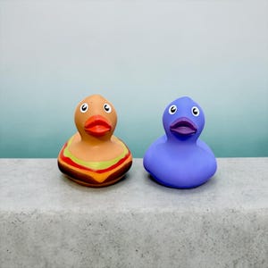 Peut inclure: Deux canards en caoutchouc. L'un est de couleur beige avec un motif de burger au milieu, l'autre est violet. Les deux ont des becs orange et des yeux noirs. Ils sont sur une surface grise avec un fond bleu et vert.