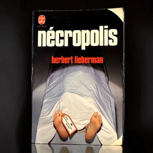 Könnte beinhalten: Buchcover von "Nécroplis" von Herbert Lieberman. Der Titel ist in großen weißen Buchstaben gehalten, der Name des Autors in Rot. Das Bild zeigt einen mit einem weißen Laken bedeckten Körper, mit freiliegenden Füßen und einem angebrachten Etikett.