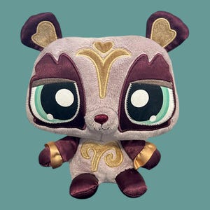 May include: A plush toy panda with a light purple body, burgundy accents, and gold embroidery. The panda has large, expressive eyes with teal and black details. Its ears and paws are burgundy with gold trim, and it has a small, embroidered nose.