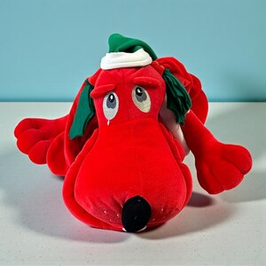 May include: A plush red dog with large, sad eyes, wearing a green and white hat. The dog has long, floppy ears and a black nose. The toy is made of soft material and is likely a children's toy or collectible.