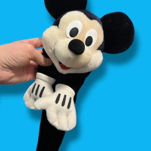 May include: A plush Mickey Mouse toy with large black ears, a black nose, and white gloves with black lines. The toy has a black body and a cream-colored face. The background is a solid blue.