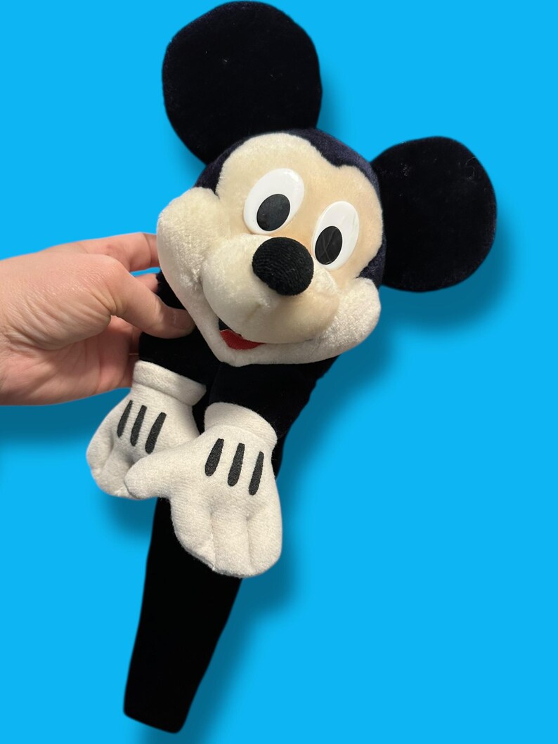 May include: A plush Mickey Mouse toy with large black ears, a black nose, and white gloves with black lines. The toy has a black body and a cream-colored face. The background is a solid blue.