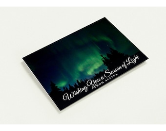 May include: A rectangular greeting card featuring a dark night sky filled with vibrant green aurora borealis lights above a silhouette of pine trees. The white cursive text reads, "Wishing You a Season of Light FROM ALASKA."