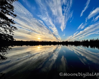 May include: A sunset over a calm lake with a dark silhouette of a forest along the horizon. Wispy, streaked clouds radiate across a bright blue sky, reflecting clearly in the still water. The text MooreDigitalAK appears in the corner.