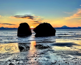 May include: Two large, dark, silhouetted rocks stand in the shallow water of a beach at sunset. The sky is filled with vibrant orange, yellow, and blue hues, reflecting on the wet sand. A distant mountain peak is visible on the horizon.