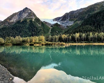 May include: A turquoise glacial lake reflects the surrounding landscape, featuring a prominent mountain peak, a distant glacier, and a dense forest of green and yellow trees. The text MooreDigitalAK is visible in the bottom right corner.