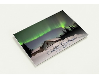 May include: A rectangular greeting card featuring a snowy landscape with a house under a vibrant green aurora borealis. The text Season's Greetings FROM ALASKA is printed in dark blue script and block lettering on the lower right side.