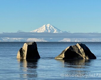 May include: Two large, dark, jagged rocks emerge from calm blue water. In the distance, a snow-capped mountain peak rises above a low layer of clouds against a clear sky. The text MooreDigitalAK is visible in the bottom right corner.