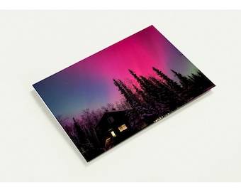 May include: A rectangular print featuring a vibrant magenta and purple aurora borealis display above a dark silhouette of a cabin nestled among tall pine trees, with warm yellow light glowing from the cabin windows against the night sky.