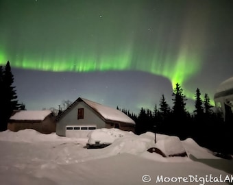 May include: A vibrant green aurora borealis stretches across a dark night sky above a snow-covered landscape featuring two buildings and silhouetted pine trees. The text MooreDigitalAK is visible in the bottom right corner of the image.