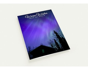 May include: A rectangular greeting card featuring a vibrant purple and blue aurora borealis sky above a dark silhouette of a cabin and pine trees. The text Warm Wishes FROM THE NORTH is printed in white script and block lettering at the top.