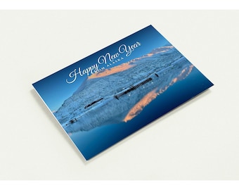 May include: A rectangular greeting card featuring a landscape photograph of a snow-covered mountain range reflected in a dark blue lake at sunset. The text "Happy New Year FROM ALASKA" is printed in white script and block lettering.