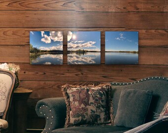 May include: A triptych wall art set featuring three square landscape photographs of a calm lake under a bright blue sky with scattered white clouds. The images show a reflection of the sky and trees on the water surface, creating a continuous horizon line across the three separate panels.