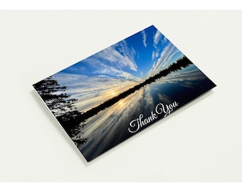 May include: A rectangular greeting card featuring a landscape photograph of a sunset over a calm lake with silhouetted trees along the shoreline. The sky is blue with wispy clouds, and the text Thank You is printed in white script in the corner.