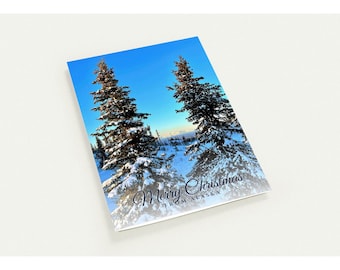 May include: A rectangular greeting card featuring a photograph of two snow-covered evergreen trees against a bright blue sky. The text Merry Christmas FROM ALASKA is printed in a dark blue script and block font at the bottom right.