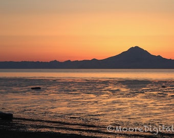 May include: A landscape view of a mountain silhouette against a vibrant orange and yellow sunset sky over a calm body of water. The foreground shows a dark shoreline with scattered rocks. The text Moore DigitalAK is visible in the corner.