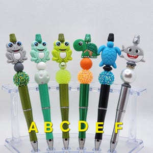 May include: Six colorful pens with different animal toppers, including a frog, turtle, and shark. The pens are displayed in a clear plastic stand labeled A through F.