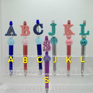 May include: A collection of decorative pens with letter and dollar sign toppers. The pens feature various colors, including blue, pink, green, and purple. Each pen has a unique design, with the letters A, B, C, J, K, L, and S displayed on the top.