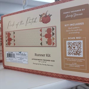 Può includere: Una scatola rettangolare per un kit runner da tavola Pick of the Patch di Sandy Gervais. La scatola presenta uno sfondo crema con bordi a quadri marroni, un'illustrazione di zucca, un codice QR e una misura di 40,6 cm per 156,2 cm.