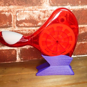 May include: A red plastic tape dispenser with a clear, rotating spool and a white handle. The dispenser is sitting on a purple stand.