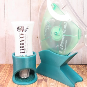 May include: A teal blue dispenser for a clear adhesive tape dispenser with the text "Scrapbook Adhesive E-Z Runner Grand Dots Repositionable" on the side.  A teal blue dispenser for a clear drying craft glue with the text "nuyo Deluxe Adhesive Clear Drying Craft Glue" on the side.