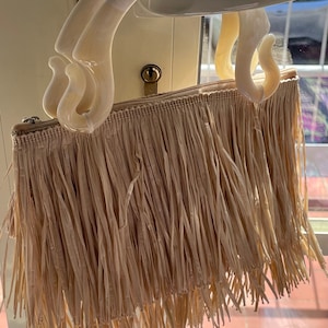 May include: A beige fringed handbag with cream-coloured, curved handles. The bag has a rectangular shape with a zip closure and is made of a woven material. The fringe hangs down from the bottom of the bag.