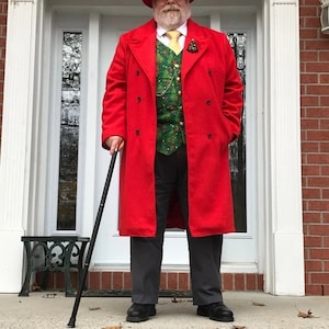 May include: A man wearing a red coat, a green vest, a gold tie, and a black hat. He is holding a black walking stick and standing in front of a door.