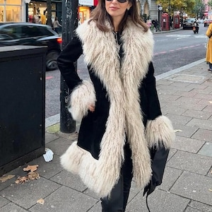 May include: A black coat featuring long sleeves and a prominent, thick, beige-colored faux fur trim along the wide lapels, the sleeve cuffs, and the bottom hemline. The coat is knee-length and has a structured fit with a smooth, dark fabric finish.