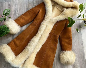 May include: A long, brown suede coat featuring a thick, cream-colored faux fur lining that extends along the front opening, the hem, and the cuffs. The coat includes a large, integrated hood lined with the same soft, cream-colored faux fur material.