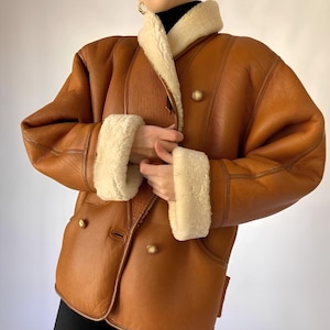 May include: A tan leather jacket featuring a cream-colored shearling collar and matching shearling cuffs. The jacket has a double-breasted front closure with large, round, gold-toned buttons and visible seam detailing throughout the garment.