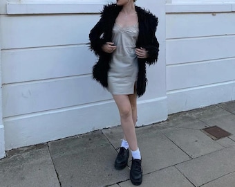Könnte beinhalten: Ein schwarzer Kunstfellmantel über einem hellfarbenen Slip-Kleid. Das Kleid hat eine Spitzenborte am Ausschnitt. Die Person trägt schwarze Plateauschuhe und weiße Socken. Der Hintergrund ist eine weiße Wand und graues Pflaster.