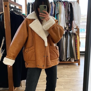 May include: A brown leather jacket with a white shearling collar and lining. The jacket has a zipper closure and two pockets on the front.