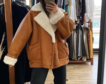 May include: A brown leather jacket with a white shearling collar and lining. The jacket has a zipper closure and two pockets on the front.
