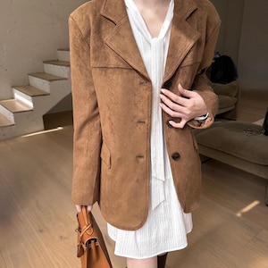 May include: A brown suede-textured blazer with a notched lapel and three front buttons, worn over a white sleeveless dress featuring a tie-neck detail. A structured brown leather handbag with a top handle is held at the side.