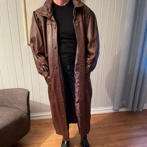 May include: A long brown leather coat with a button closure and a collar. The coat has a slightly flared shape and is lined with a purple fabric.