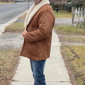 May include: A brown suede coat with a cream-colored shearling collar. The coat has a classic design with large pockets and a button closure. The person is wearing blue jeans. The coat appears to be warm and suitable for cold weather.