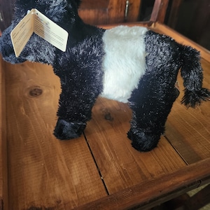 Op de afbeelding: Zwart-witte pluche koe met een witte vlek aan de zijkant. De koe staat op een houten oppervlak. Het label op de koe luidt "Little Adventures Plush Cow".