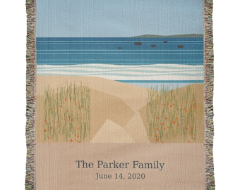 May include: A woven throw blanket featuring a beach scene with sand dunes, tall grass, and a blue ocean under a light sky. The bottom section is tan and displays the text The Parker Family and the date June 14, 2020 in dark grey lettering.